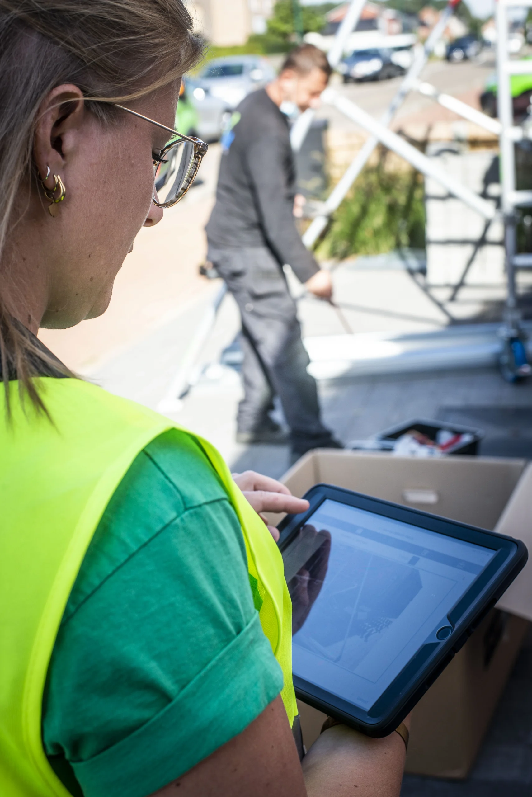 Een dame met een groen T-shirt en fluohesje staat op een werf te kijken naar haar tablet waarop een foto van de werf staat.