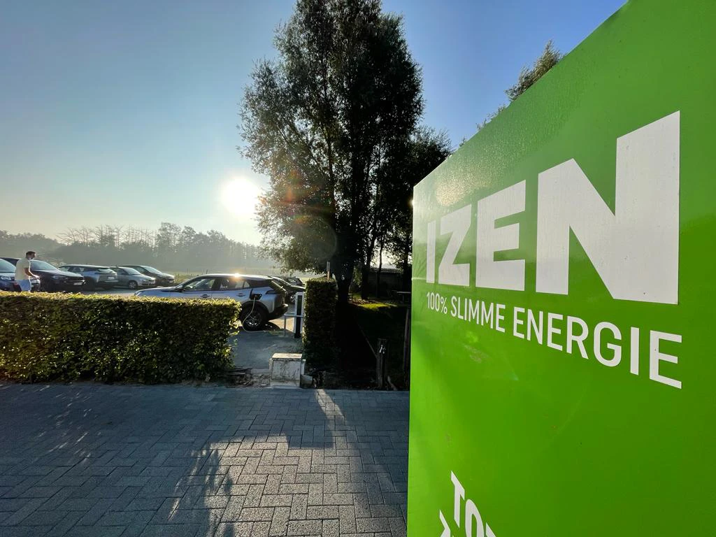 De parking bij het kantoor van IZEN. Op de parking staan een aantal wagen waaronder ook een elektrische auto is ingeplugd is in een laadpaal. In de achtergrond zijn open, groene velden. In de voorgrond staat een groen bord met het logo van IZEN in wit.