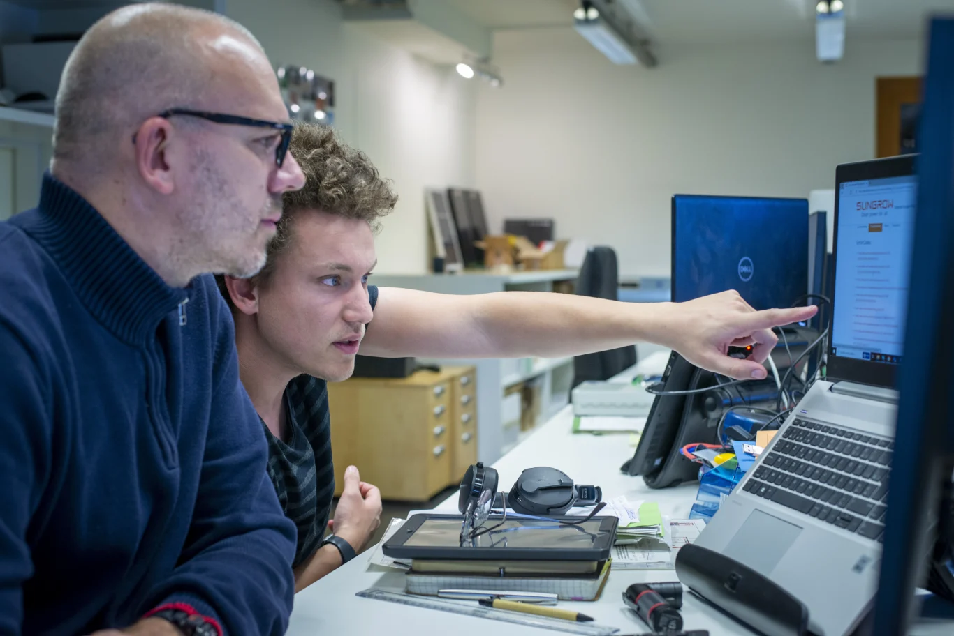 2 medewerkers van IZEN kijken samen naar een computerscherm. Eén van de mannen wijst naar het scherm waarop een webpagina te zien is van omvormer-fabrikant Sungrow